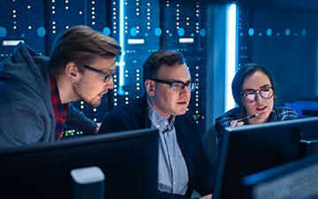 Two men and a woman, all wearing glasses, utilizing Viasat software on computers in a server room