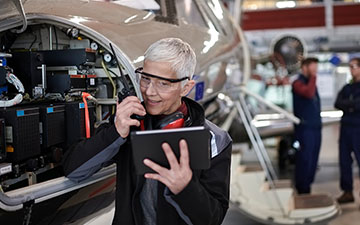 Female airplane technician wearing safety glasses, talking into a radio while looking at a tablet next to an airplane