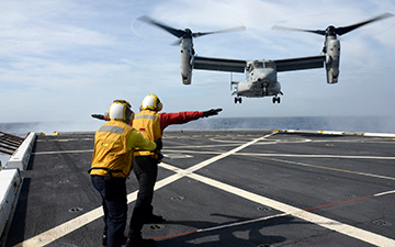 Two men wearing yellow vests helping a military plane land on a ship at sea